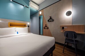 Habitación de hotel con cama, escritorio y silla. Se observa una unidad de aire acondicionado en la pared y una puerta con picaporte.