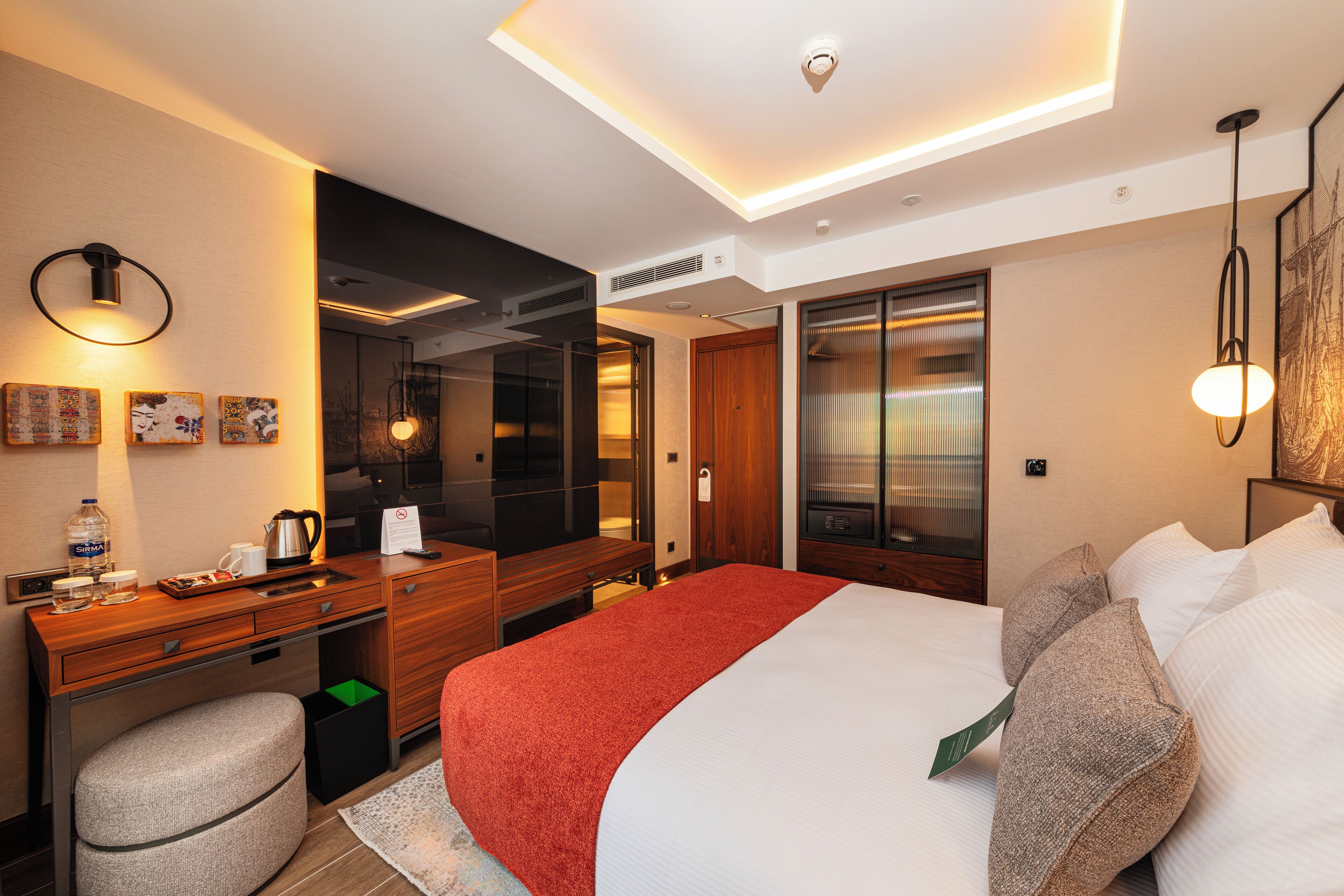 A modern hotel room with a large bed, a desk with a chair, a mirror, and a wardrobe.