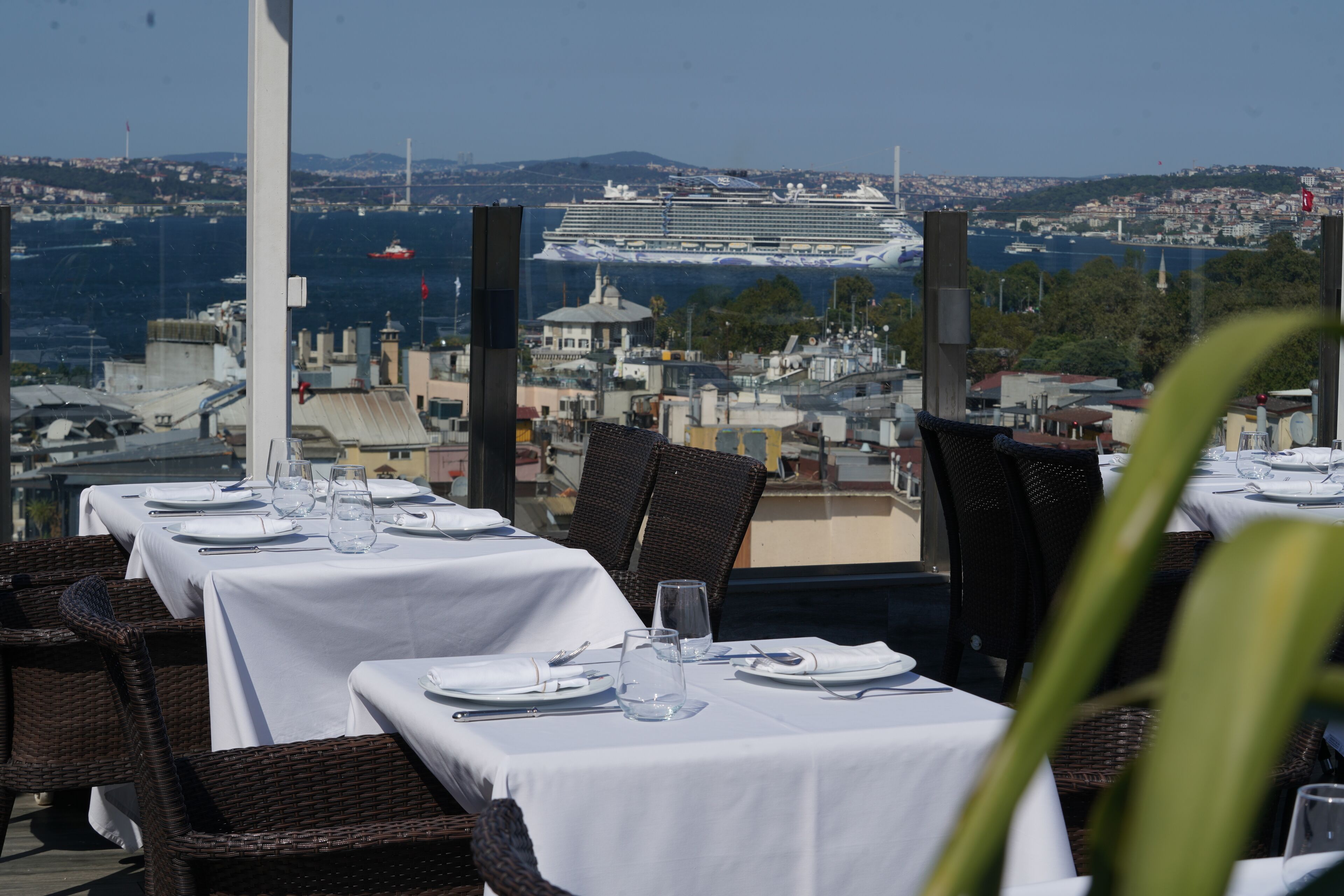 Ein Restaurant mit gedeckten Tischen, mit Blick auf eine Stadtlandschaft und ein Kreuzfahrtschiff in der Ferne.