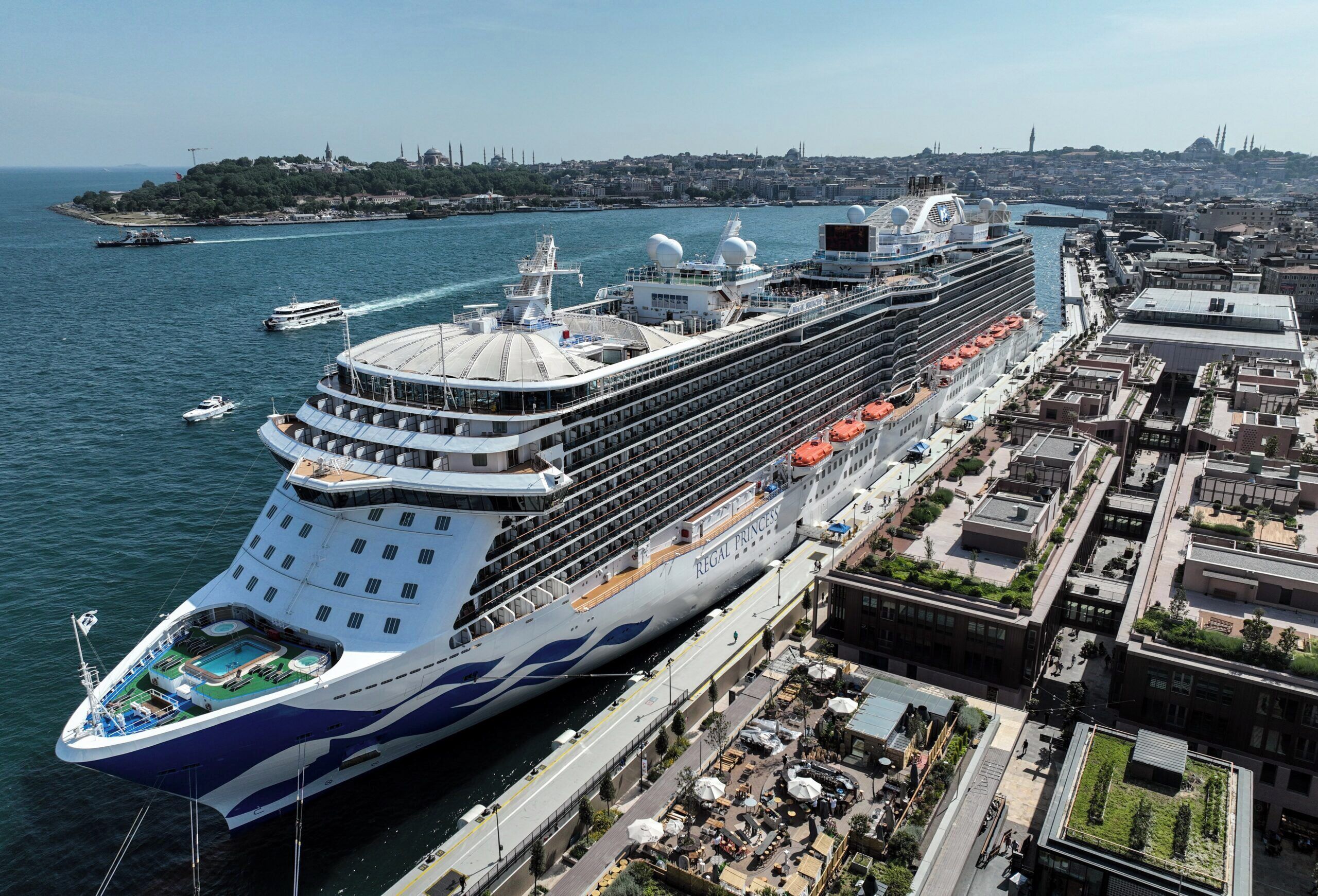 A large cruise ship docked at a port with multiple decks and a cityscape in the background.