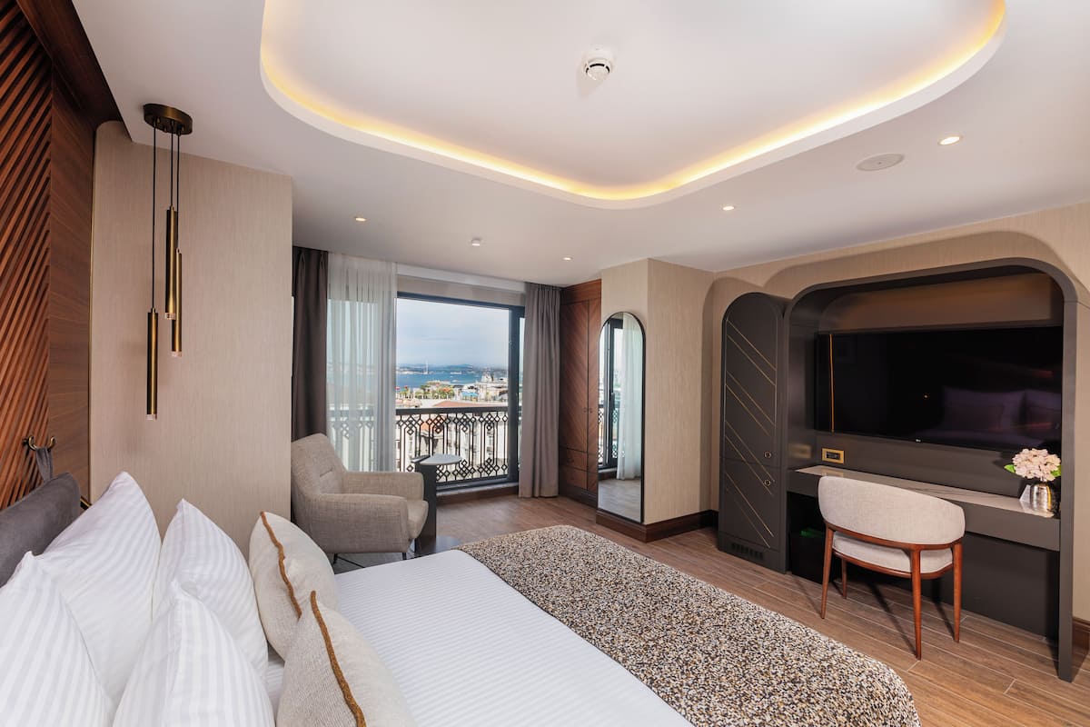 A modern hotel room with a large bed, a desk with a chair, a TV, and a balcony with a city view.
