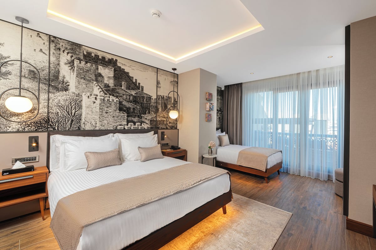 A modern hotel room with a large bed, a smaller bed, a nightstand with a lamp, a wall-mounted TV, and a large window with sheer curtains.