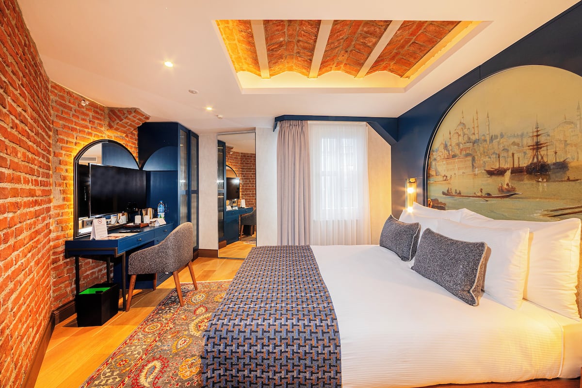 A hotel room with a brick wall, a large bed with white linens, a desk with a computer, and a painting of a cityscape on the wall.