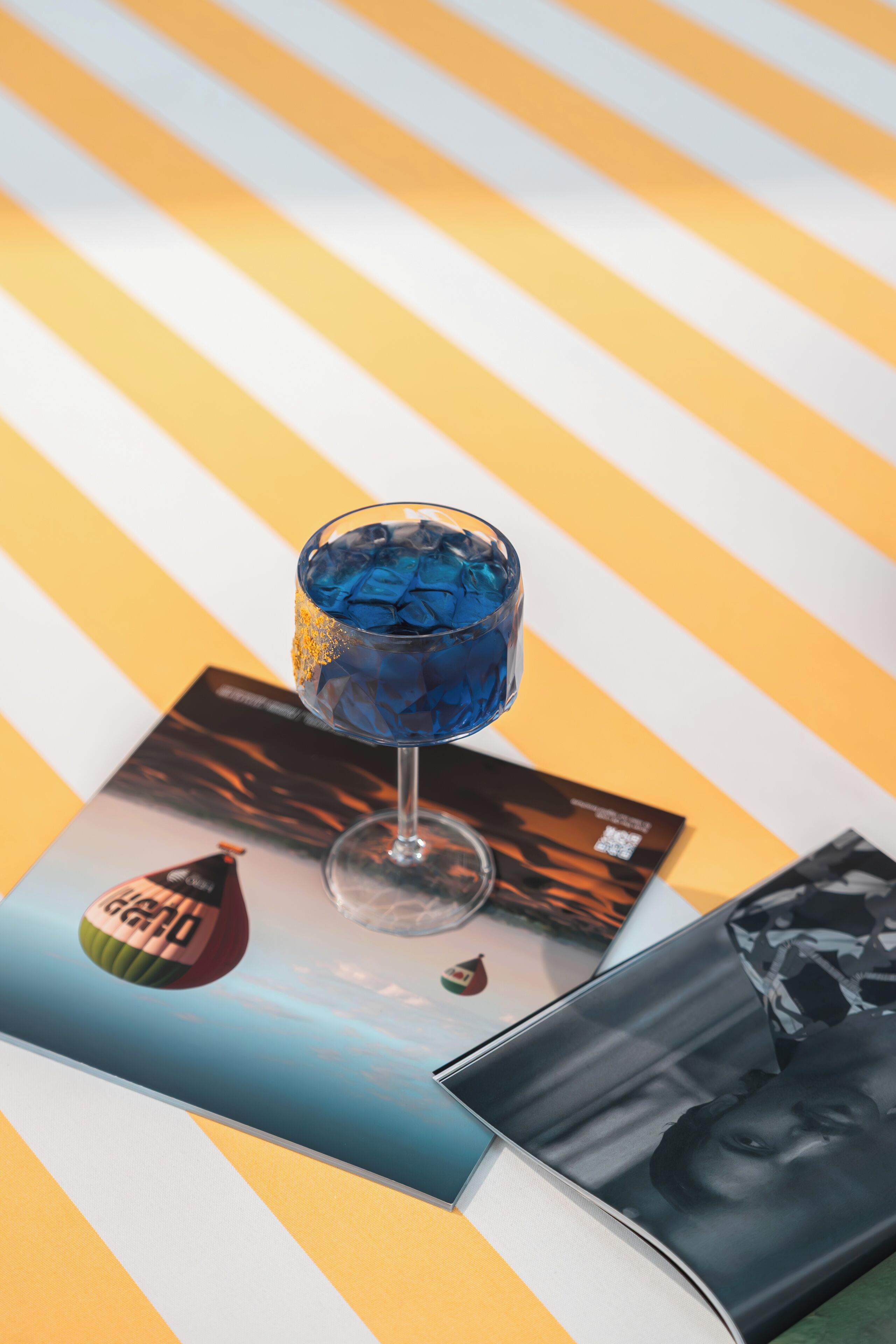 Ein Glas mit blauen Eiswürfeln auf einer gestreiften Fläche.