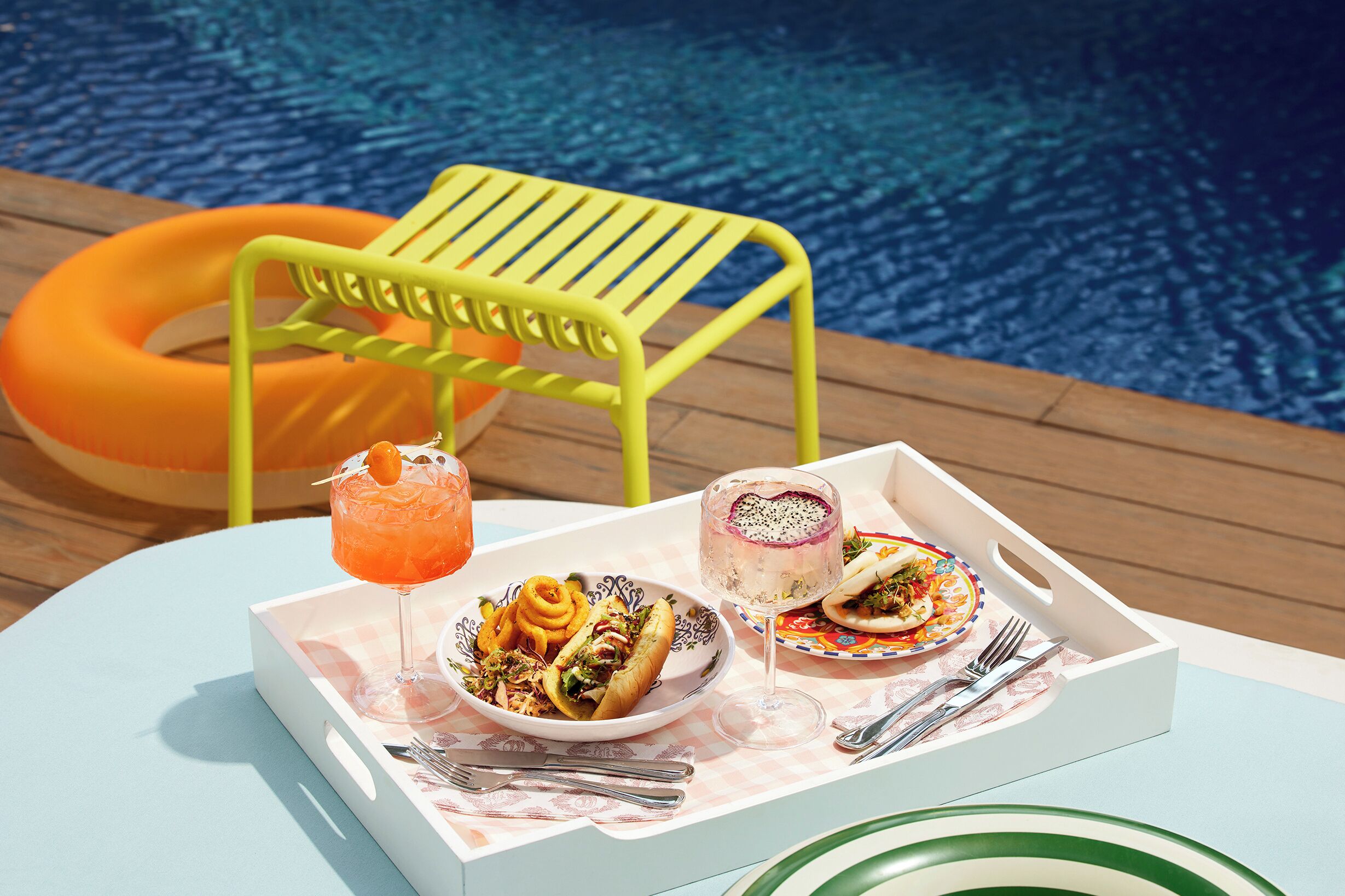 A dining table with a tray of food and drinks, a yellow chair, and a pool in the background.