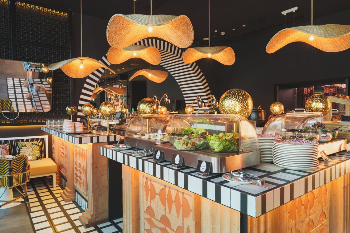 A buffet area with a variety of food items, a seating area with chairs, and a unique ceiling design with hanging light fixtures.