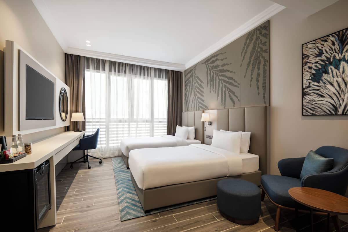 A hotel room with two beds, a desk with a TV, a chair, a small table, and a large window with curtains.