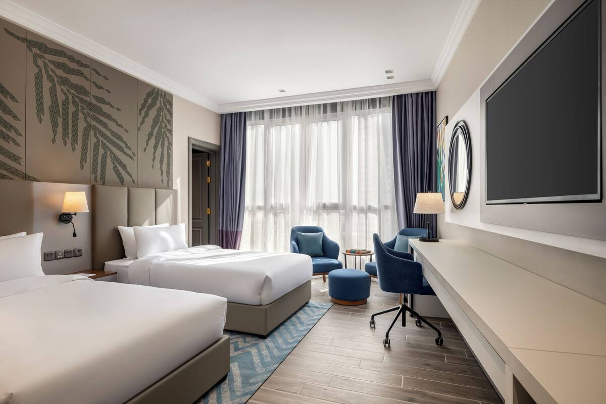 A hotel room with two beds, a desk, a chair, a TV, and a large window with curtains.