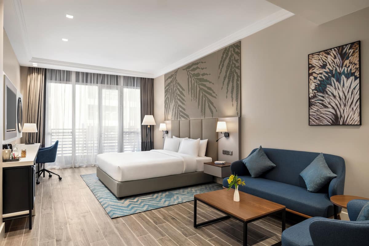 A modern hotel room with a large bed, a sofa, a desk with a chair, and a coffee table.