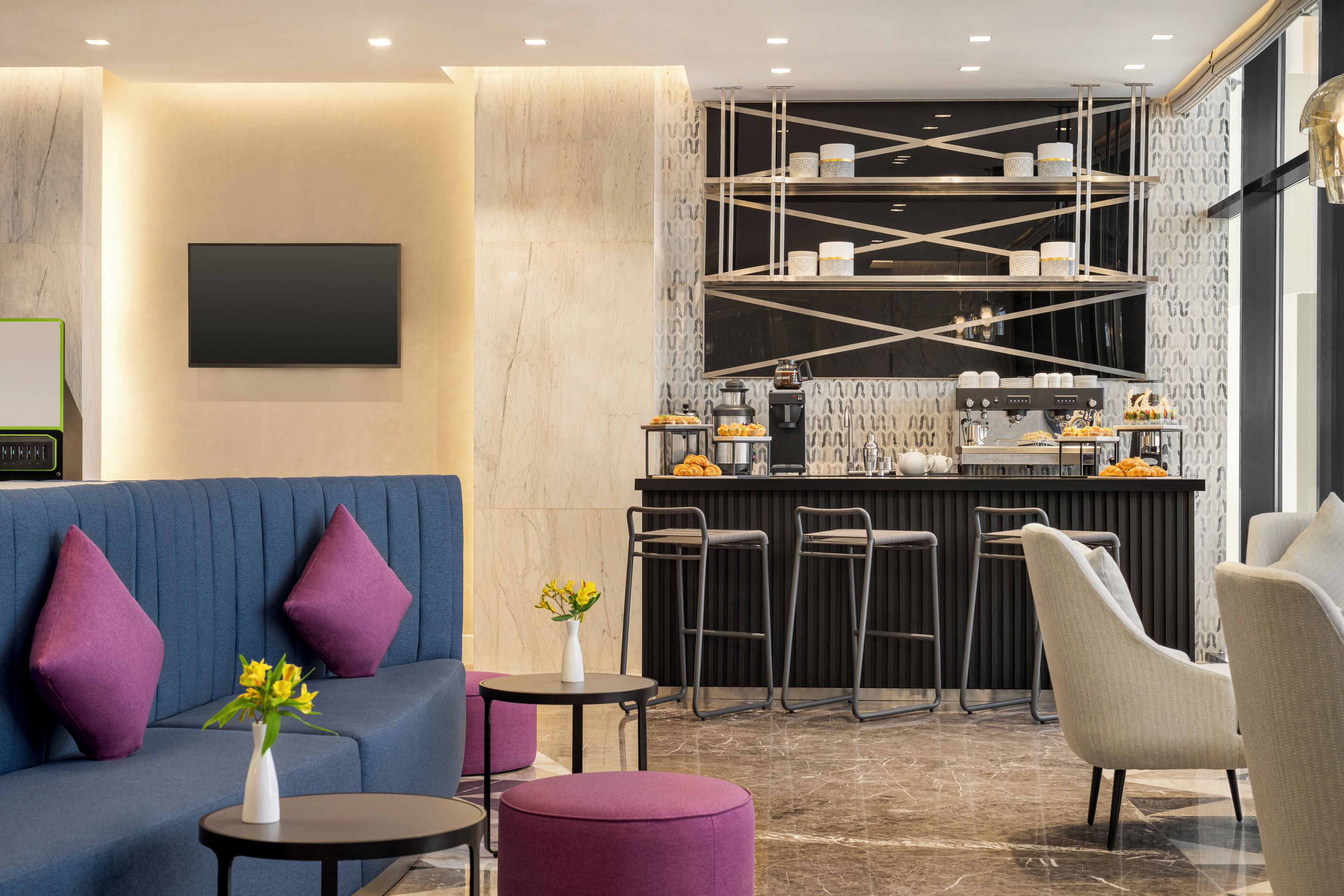 A modern hotel lobby with a blue sofa, purple cushions, a small round table with a vase, and a bar area with stools and kitchen appliances.