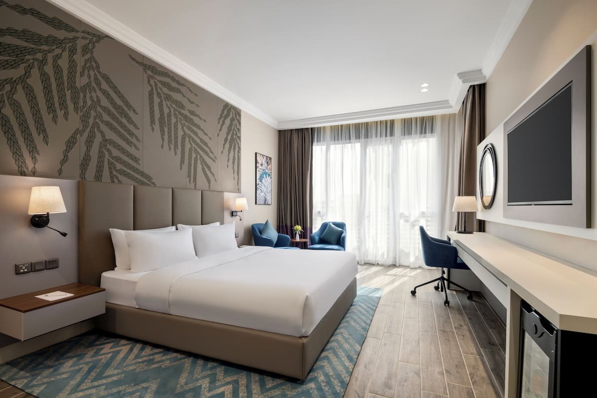 A modern hotel room with a large bed, two bedside tables with lamps, a desk with a chair, and a large window with curtains.