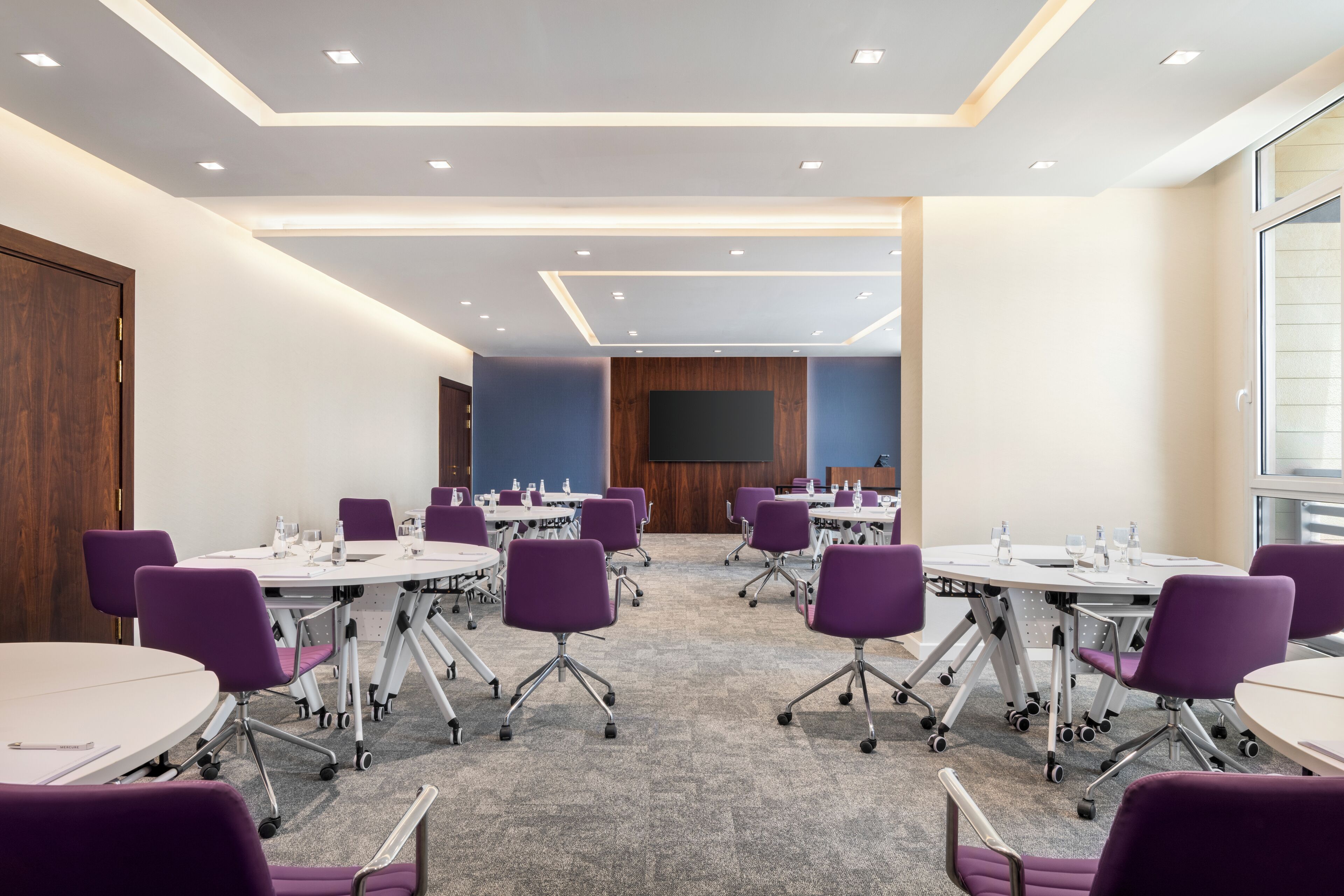A conference room with round tables, purple chairs, and a large screen at the front.