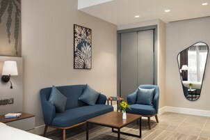 A modern hotel room with a sofa, armchair, and a coffee table. There is a painting on the wall, a mirror, and a small table with a vase of flowers.