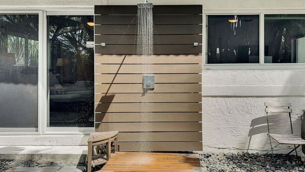 A modern outdoor shower area with a wooden bench, a chair, and a stone pathway. - Towering Palms in Wilton Manors (Wilton Manors)