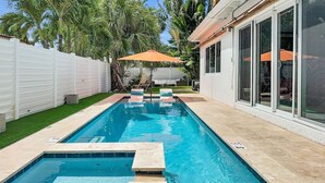 A rectangular swimming pool with clear blue water, surrounded by a tiled deck. There are two lounge chairs with an orange umbrella, and a white fence enclosing the area. - Towering Palms in Wilton Manors (Wilton Manors)