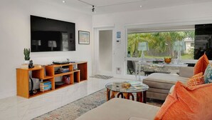 A modern living room with a large flat-screen TV, a wooden bookshelf, a beige sofa, and a glass coffee table. - Towering Palms in Wilton Manors (Wilton Manors)
