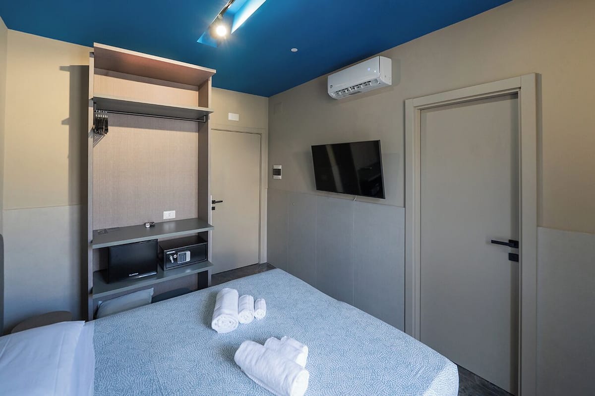 A hotel room with a bed, a TV mounted on the wall, a wardrobe, and an air conditioning unit.
