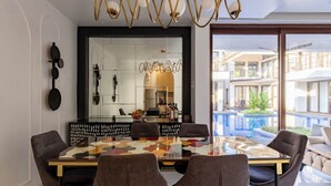 A modern dining area with a glass table, six chairs, a decorative mirror, and a view of a swimming pool and buildings. - Casa Amore Aqua Sol (Vagator)