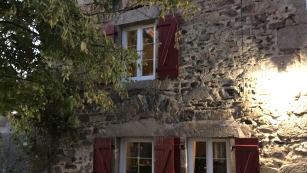 Ein Steinhaus mit weißer Tür und roten Fensterläden, mit einer kleinen Veranda und Kübelpflanzen.