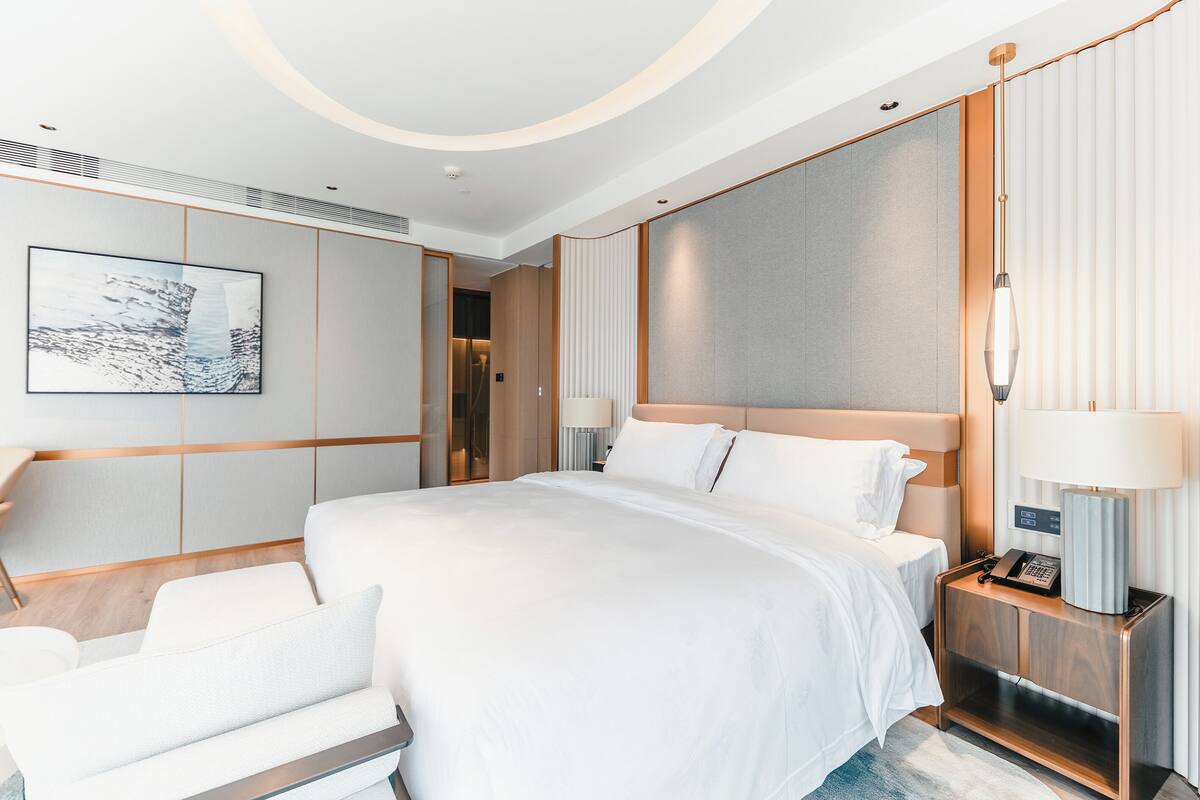 A modern hotel room with a large bed, a bedside table with a lamp, a chair, and a wall-mounted artwork.