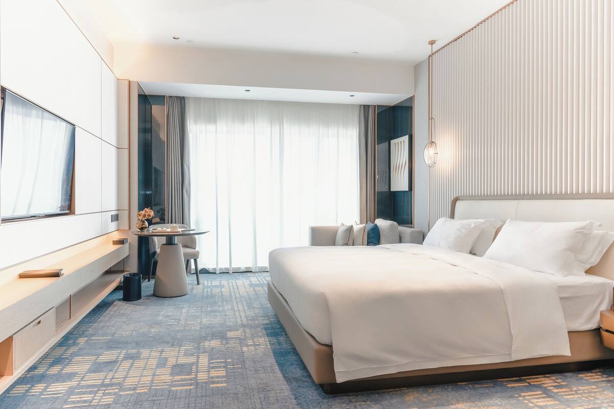 A modern hotel room with a large bed, a desk with a chair, and a view of the city through large windows.