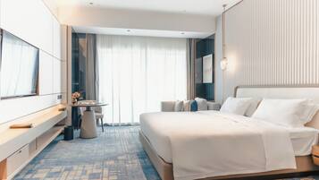 A modern hotel room with a large bed, a desk with a chair, and a view of the city through large windows.