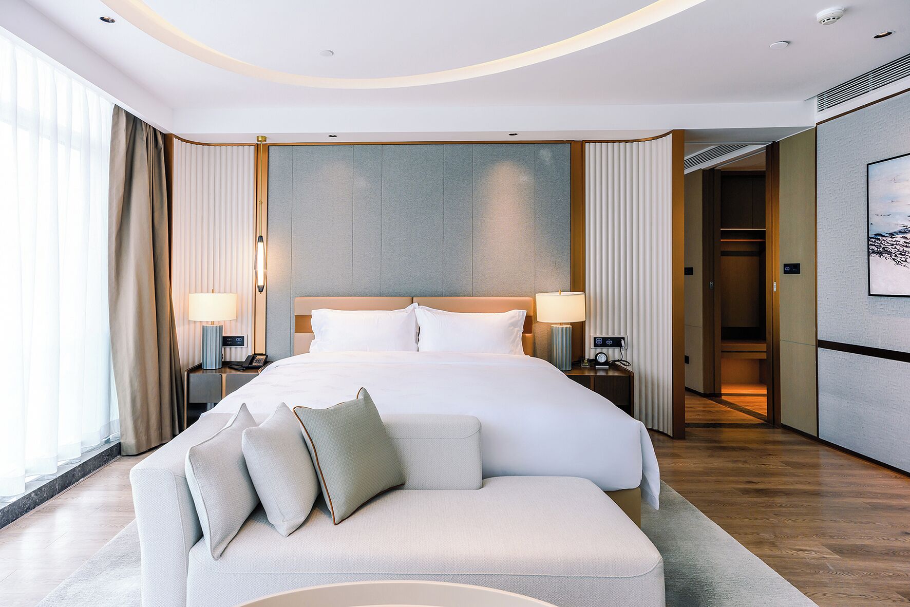Ein modernes Hotelzimmer mit einem großen Bett, zwei Nachttischen mit Lampen, einer Couch mit Kissen und einem großen Fenster mit Vorhängen.