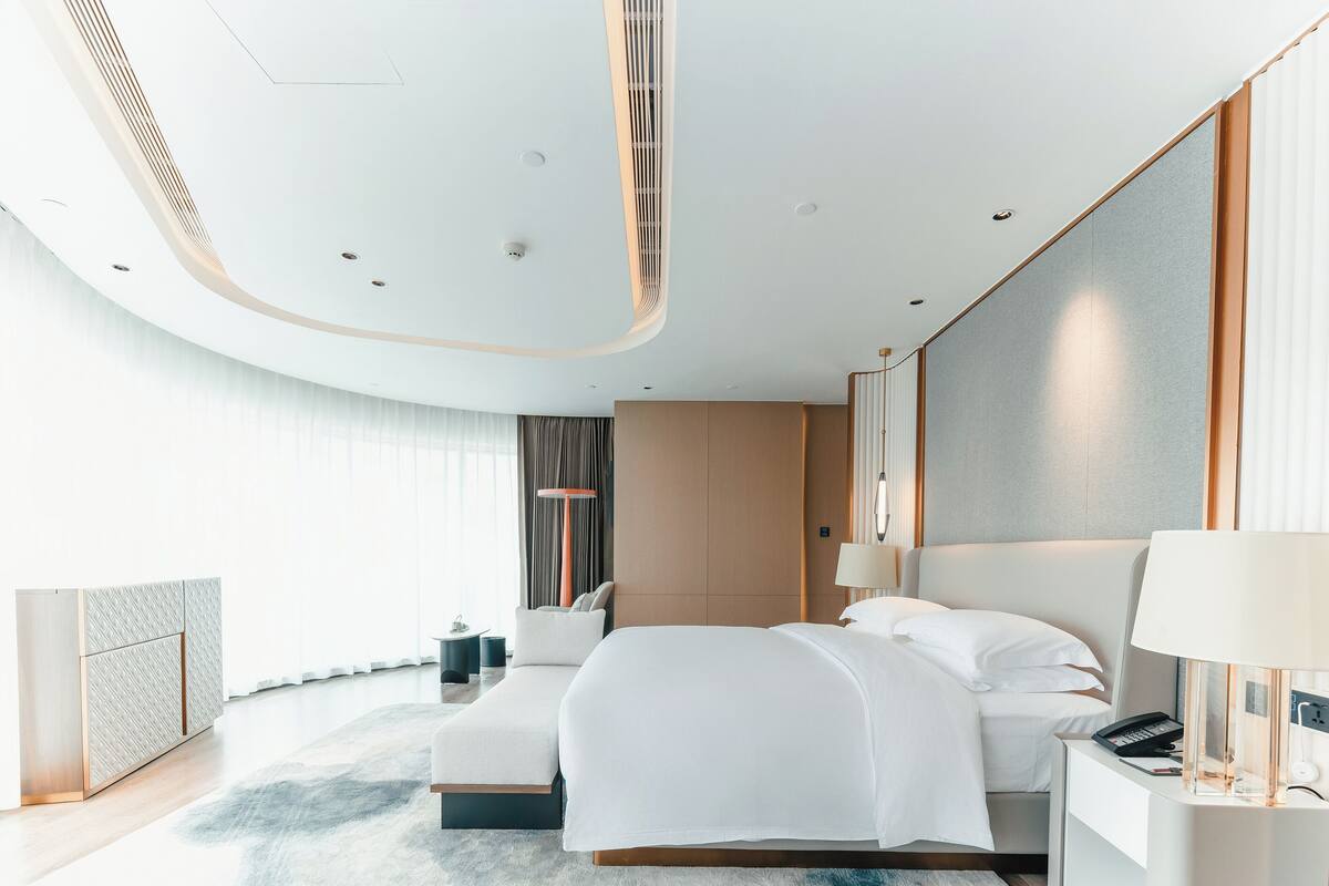 A modern hotel room with a curved ceiling, a large bed, a bedside table with a lamp, and a seating area with a sofa and a small table.