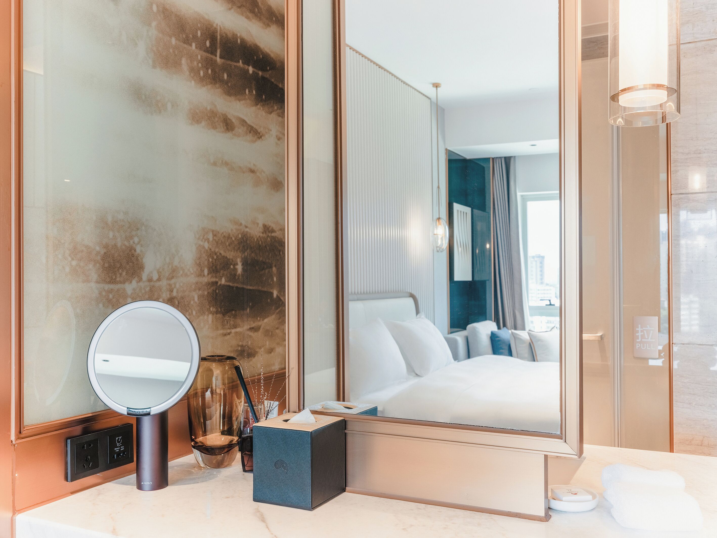 A modern hotel room with a large mirror, a bedside table with a lamp, a box, and a towel on the bed.