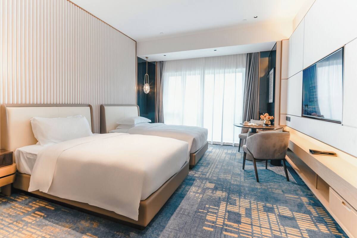 A hotel room with two beds, a desk, a chair, a TV, and a large window with curtains.