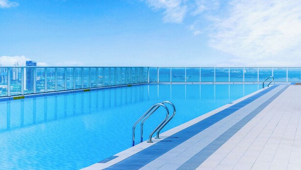 A clear blue sky with a cityscape in the background, a swimming pool with a glass barrier, and a tiled deck.