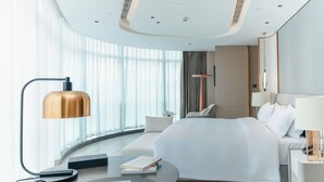 A modern hotel room with a curved bed, a bedside table with a lamp, a round coffee table, and a view of the city through large windows.