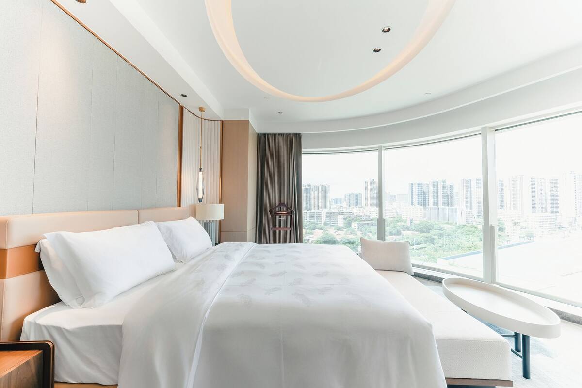 A modern hotel room with a large bed, a bedside table, a chair, and a view of the cityscape through large windows.