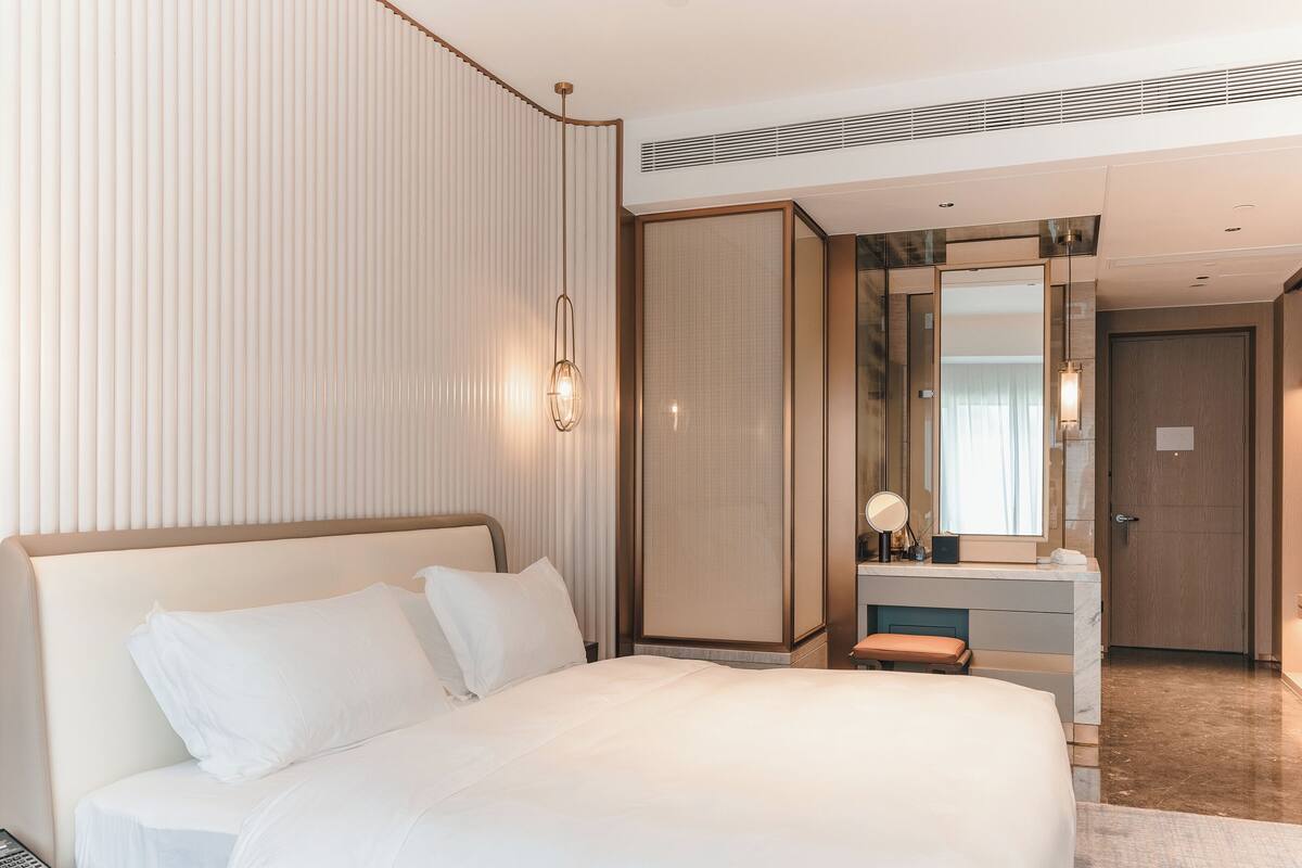 A hotel room with a large bed, a bedside table with a phone, a desk with a mirror, and a wardrobe.