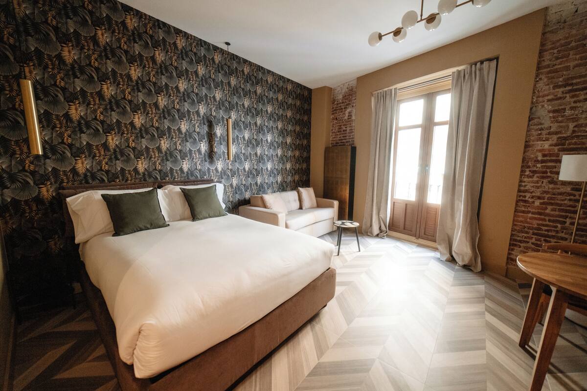 A modern hotel room with a large bed, a sofa, a small table, and a large window with curtains.