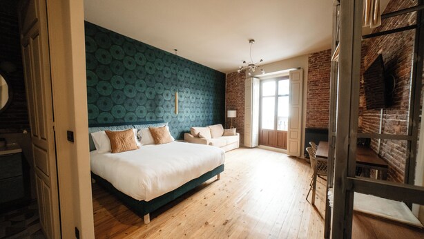 A bedroom with a large bed, a wooden floor, a blue patterned wall, and a brick fireplace. - Rincón del Conde Suites (León)