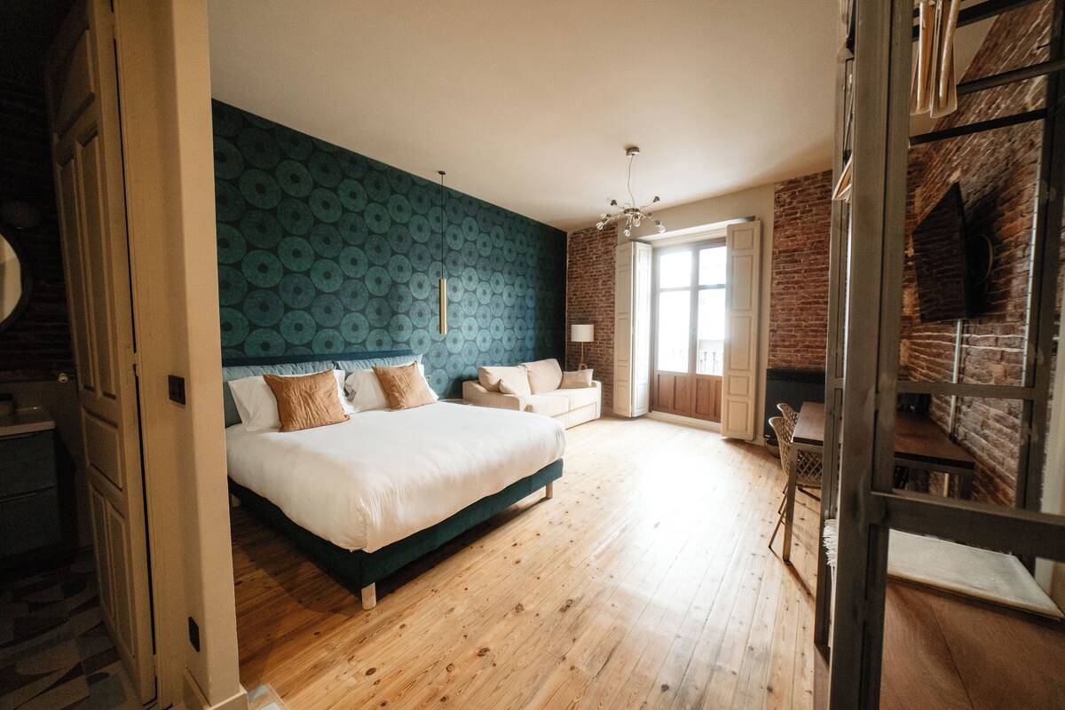 A bedroom with a large bed, a wooden floor, a blue patterned wall, and a brick fireplace.
