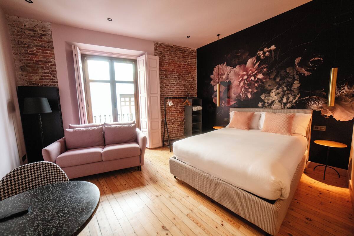 A bedroom with a large bed, a sofa, a small table, and a brick wall.