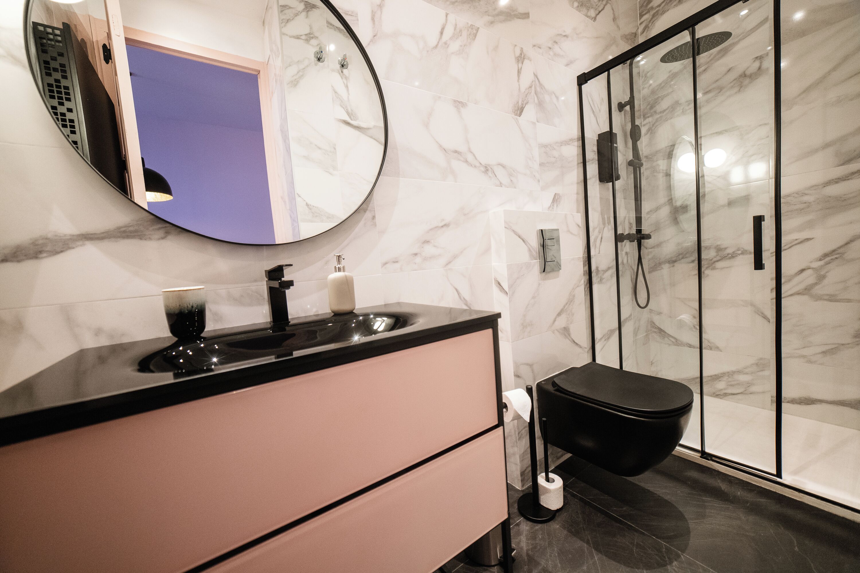 A modern bathroom with a marble wall, a round mirror, a black sink, a toilet, and a glass-enclosed shower.