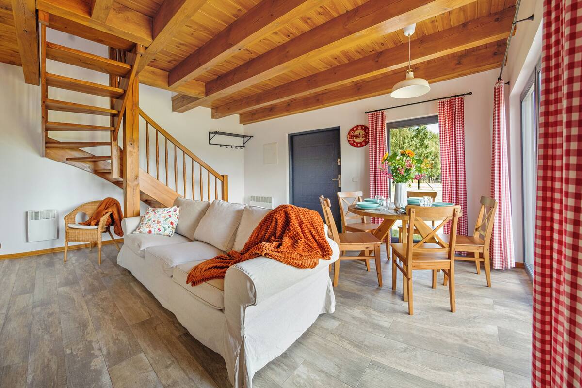 Un salon chaleureux avec un escalier en bois, un canapé, une table à manger avec des chaises et une fenêtre ornée de rideaux à carreaux.