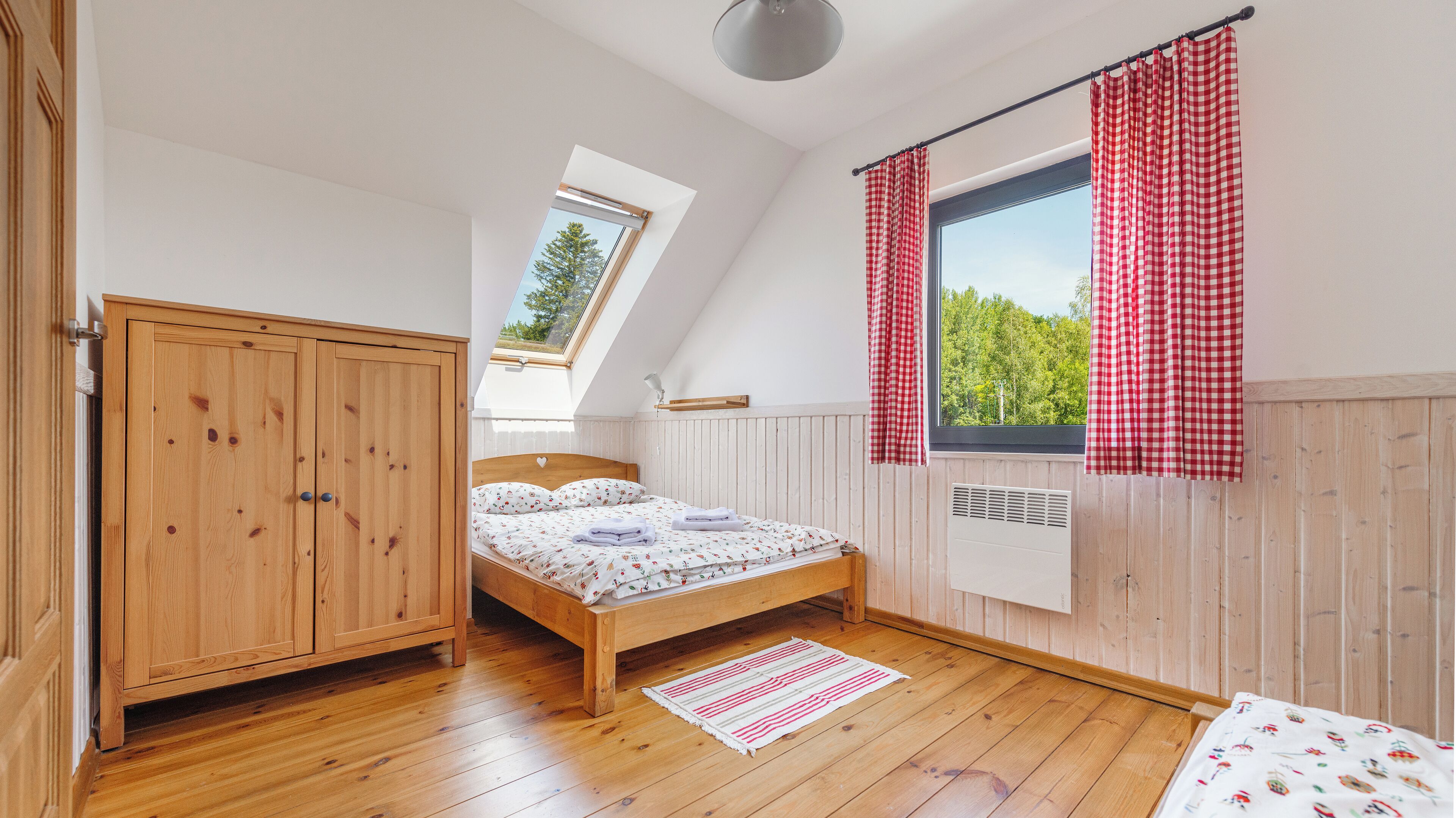 A bedroom with a wooden wardrobe, a bed with a patterned duvet, a skylight, a radiator, and a window with red checkered curtains.