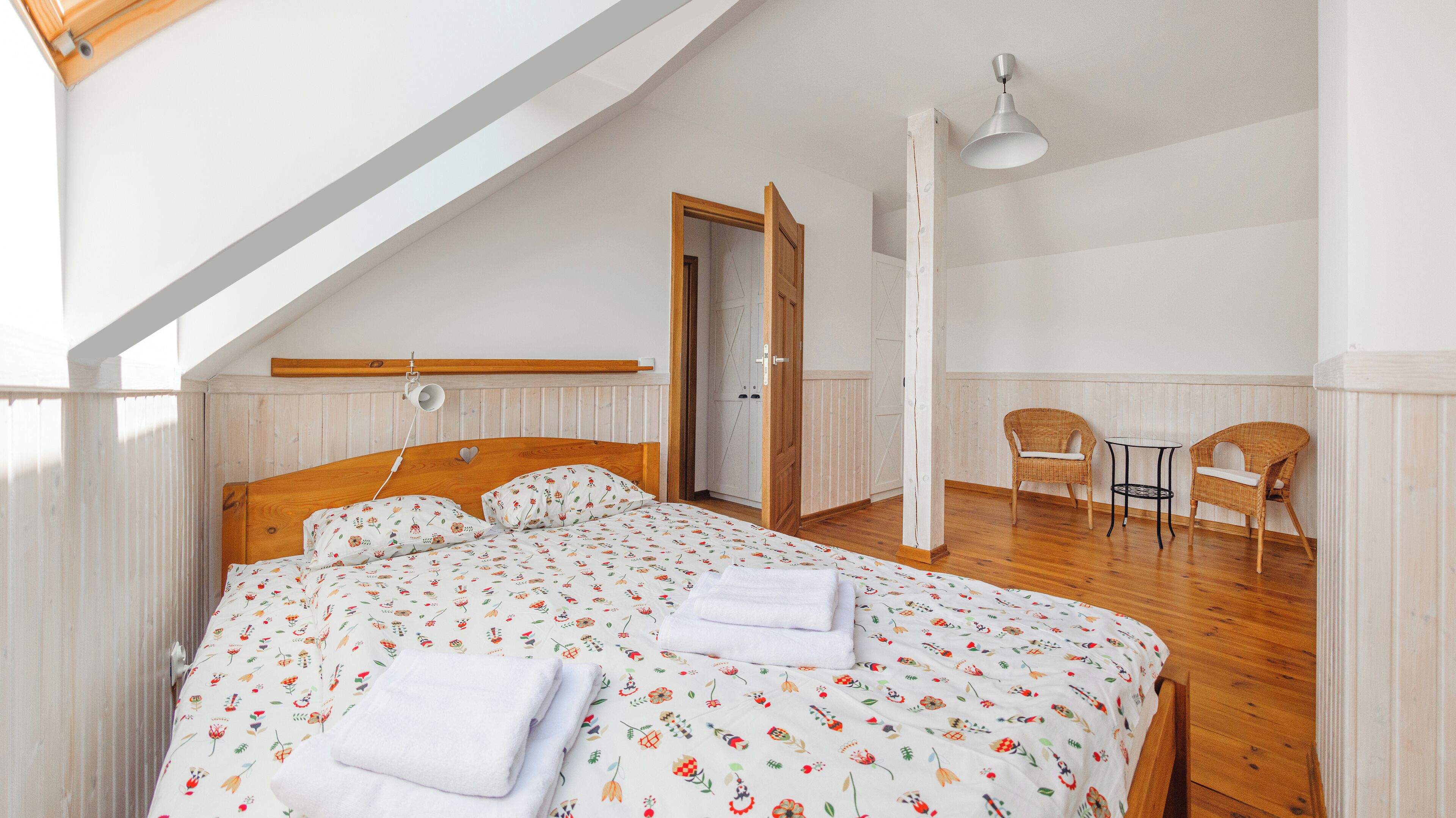 A bedroom with a wooden bed, a white wall, a wooden door, and a small table with two wicker chairs.