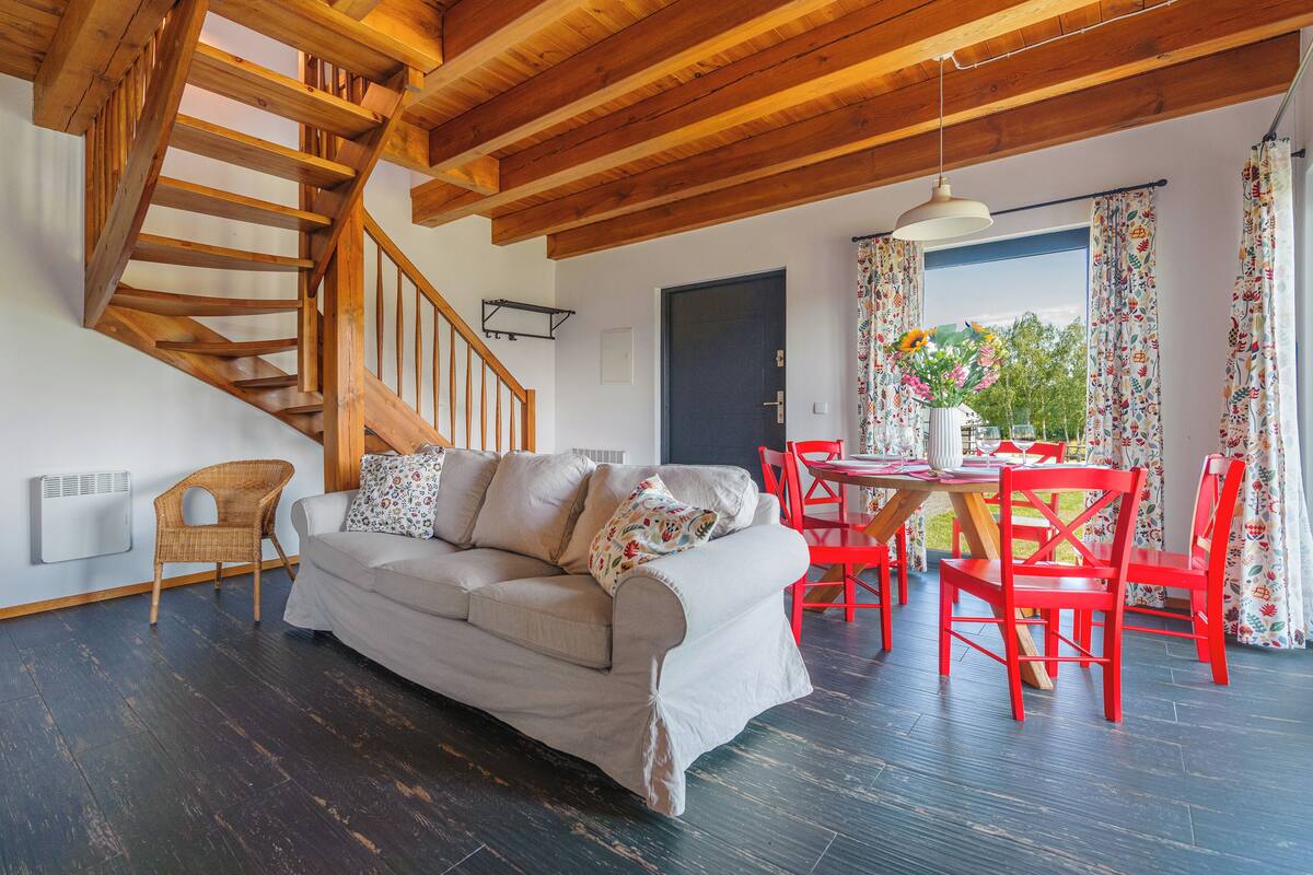 Un salon chaleureux avec un escalier en bois, un canapé, une table à manger entourée de chaises rouges et un téléviseur fixé au mur.