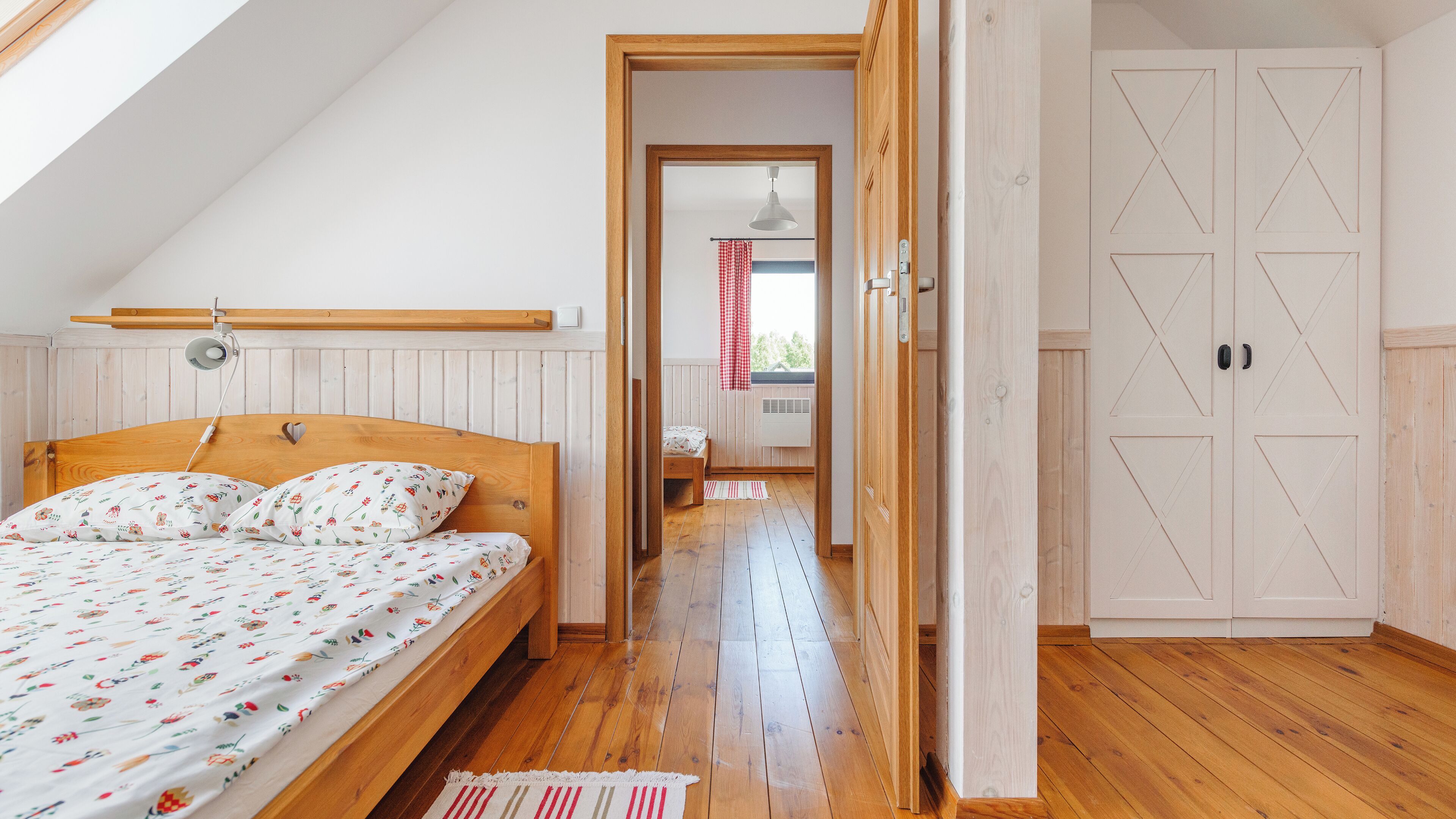 A bedroom with a wooden bed, a sloped ceiling, and a view into another room with a window and a red curtain.