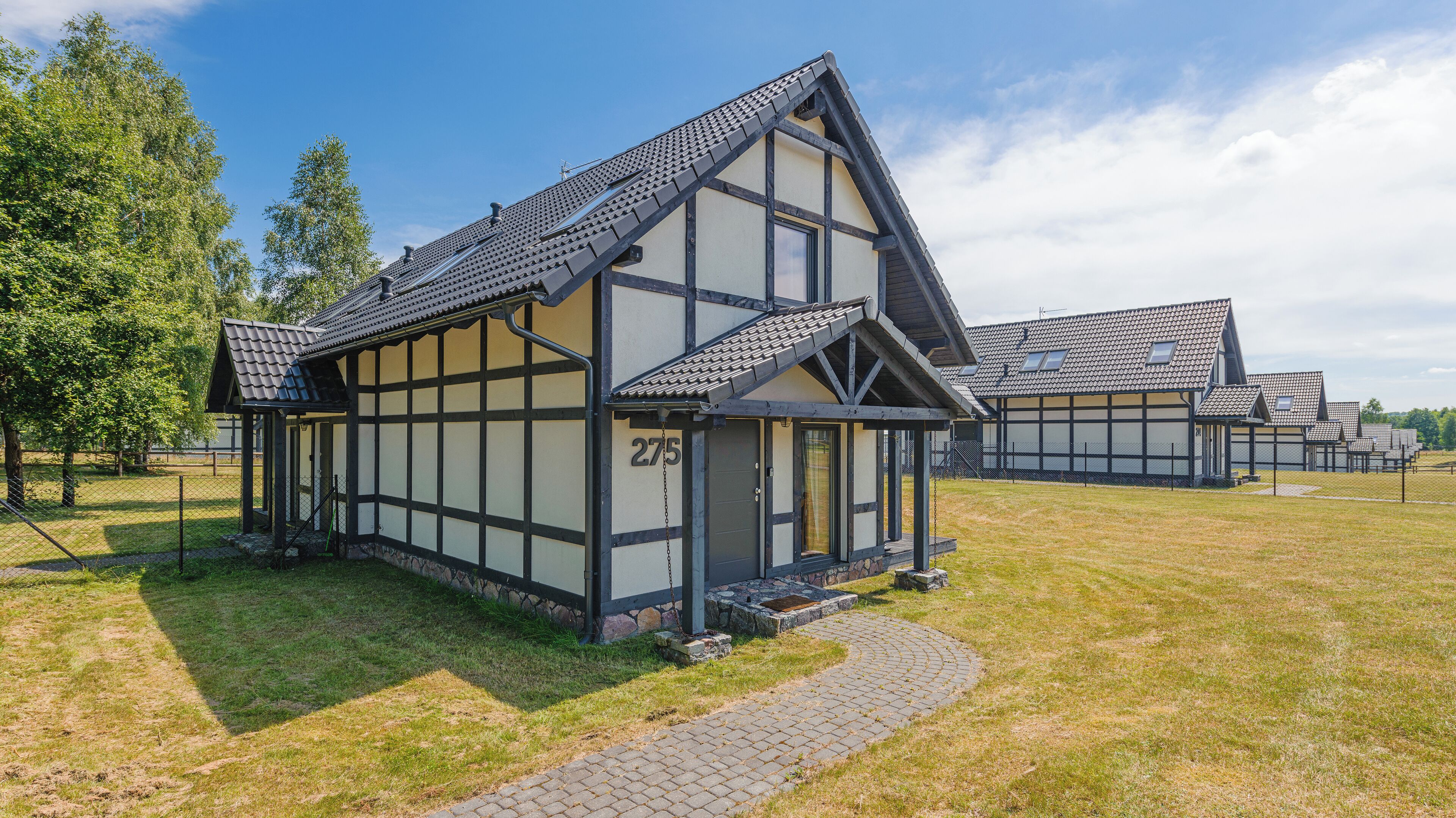 A traditional half-timbered house with a steep gabled roof, numbered 275, set in a grassy area with a paved path leading to the entrance.
