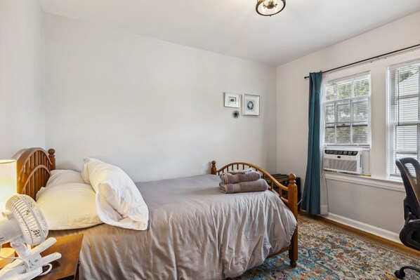 A bedroom with a bed, a bedside fan, a chair, a window with curtains, and an air conditioning unit.