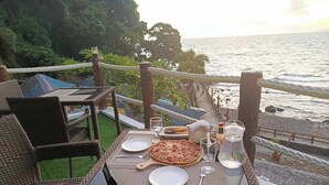 Ein Esstisch, gedeckt mit Tellern, einer Pizza, Besteck und einem Glas Wasser, mit Blick auf einen Strand und das Meer.