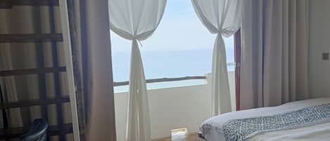 Un dormitorio con cama, piso de madera y vista al mar a través de cortinas translúcidas.