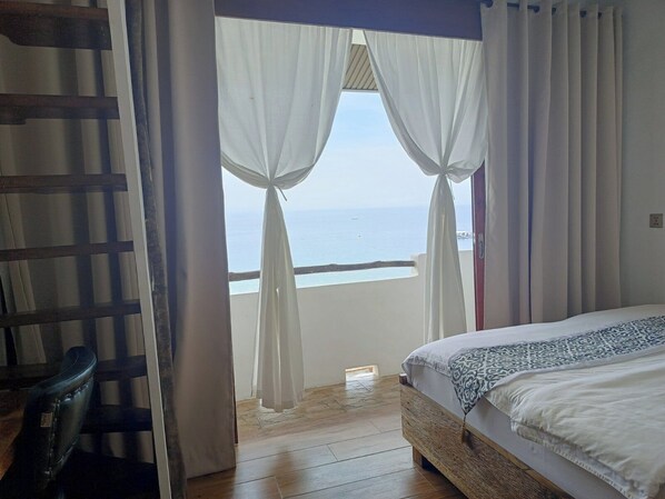 Un dormitorio con cama, piso de madera y vista al mar a través de cortinas translúcidas.