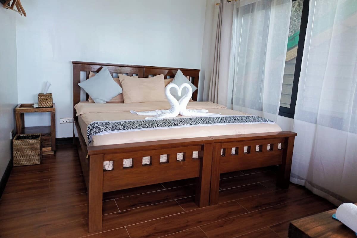 Un lit bien fait, agrémenté d’une serviette en forme de cœur, avec une tête de lit en bois et une table de chevet sur laquelle se trouve un panier.